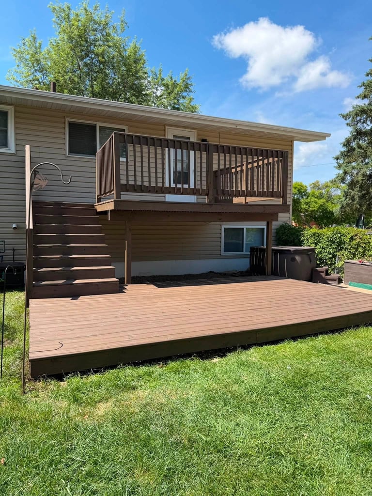 Residential house with elevated deck and railing, multi-level wooden structure, green lawn and mature trees on sunny day