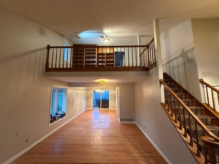 Interior of a two-story residential space with wooden flooring, loft area with built-in shelving, and staircase with wooden railings.