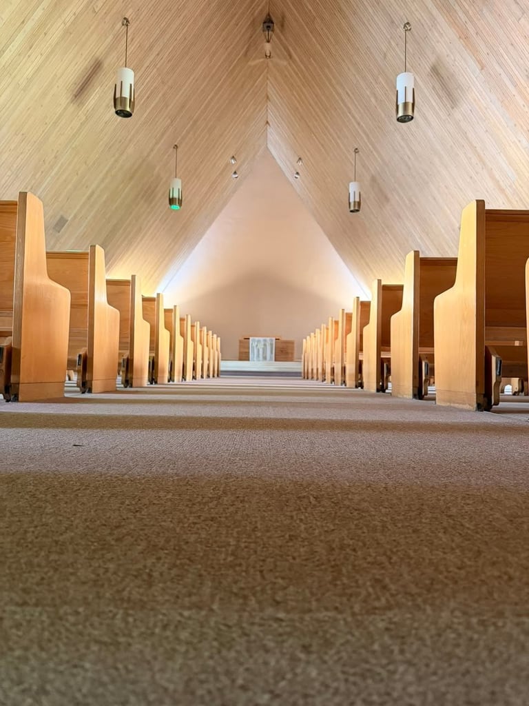 Modern church interior with symmetrical wooden pews lining a central aisle, vaulted ceiling with pendant lights, and altar at the far end