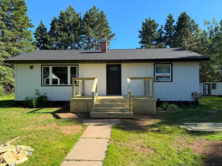 Single-story white ranch house with wooden deck, surrounded by green lawn and evergreen trees on a sunny day
