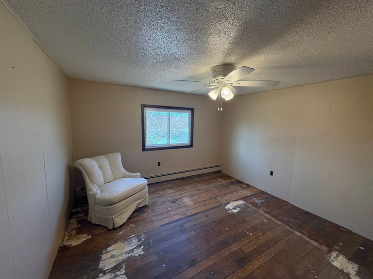 Empty room with cream-colored walls, hardwood floor, ceiling fan, window, and a single upholstered chair