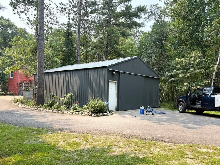 A dark metal-sided workshop or garage building with a peaked roof sits in a wooded lot, with a pickup truck parked outside