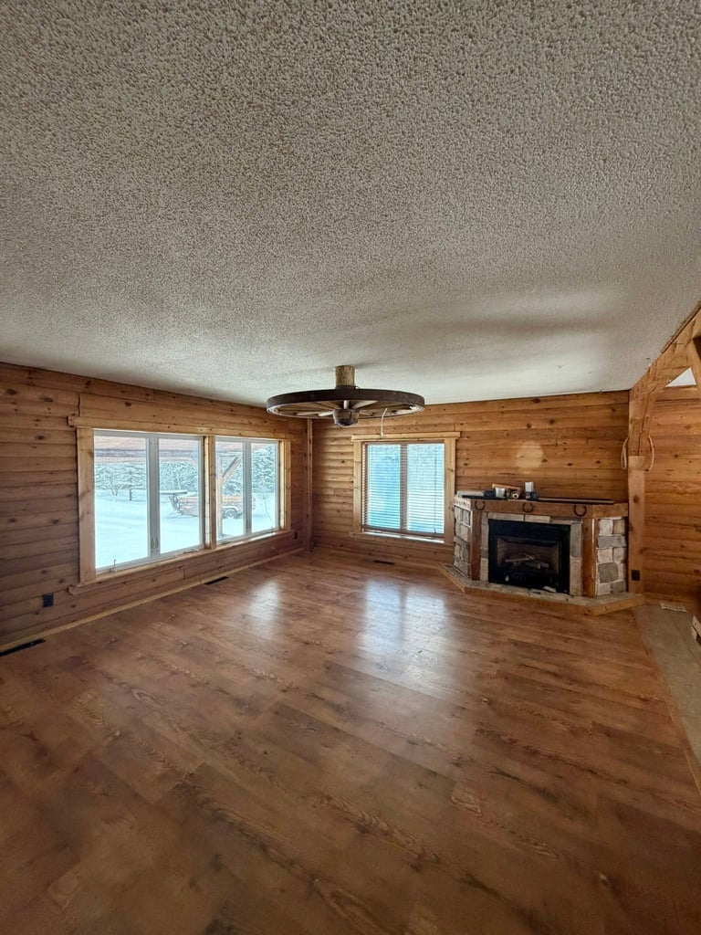 Spacious wooden log cabin interior with hardwood floors, fireplace, ceiling fan, and large windows overlooking snowy landscape