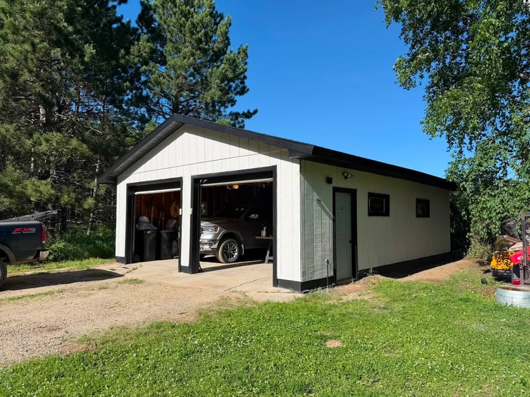 Modern white and black garage with open bay doors, motorcycle inside, surrounded by green lawn and tall pine trees on clear day